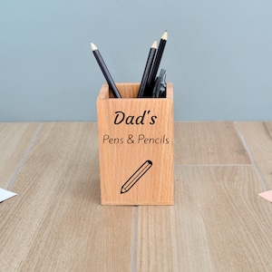 May include: A wooden pencil holder with the text "Dad's Pens & Pencils" and a pencil icon. The holder is filled with black pens and pencils.