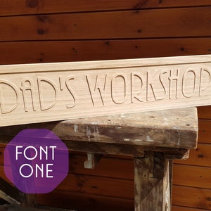 May include: A wooden sign with the text "Dad's Workshop" carved into it. The sign is light brown and has a slightly distressed look. The text is in a decorative font.