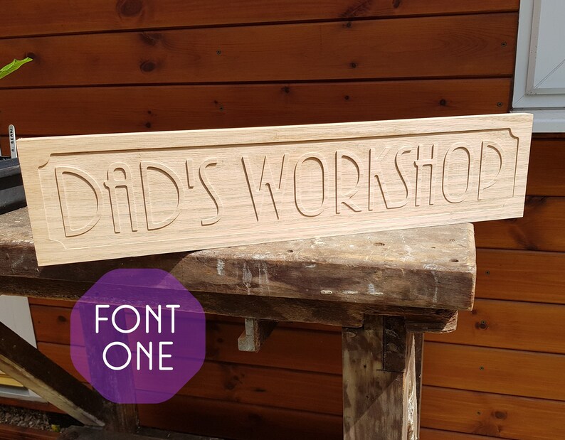 May include: A wooden sign with the text "Dad's Workshop" carved into it. The sign is light brown and has a slightly distressed look. The text is in a decorative font.