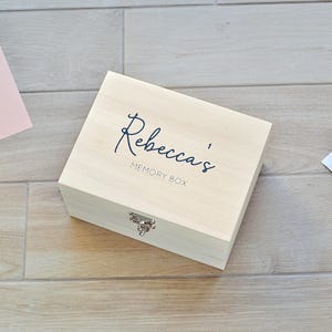 Peut inclure: Un coffret souvenir en bois clair avec le nom "Rebecca's" en écriture cursive sur le couvercle. La boîte a un fermoir argenté et est posée sur une surface en bois. Les mots "MEMORY BOX" sont en dessous.