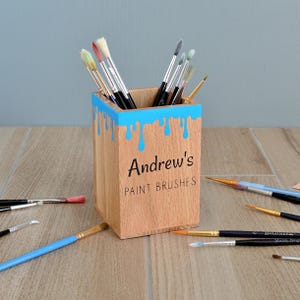May include: Wooden paintbrush holder with a blue paint drip design. The holder is personalised with the name "Andrew's Paint Brushes".