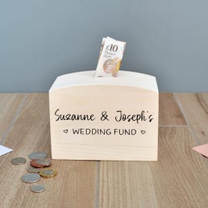 Può includere: Un salvadanaio di legno con uno slot in cima per i soldi. Il salvadanaio è inciso con il testo "Suzanne & Joseph's WEDDING FUND".