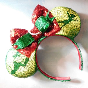 Puede incluir: Una diadema festiva con orejas de lentejuelas y un lazo grande. Las orejas son verdes y doradas, con la silueta de un personaje. El lazo es rojo y verde, con un acento decorativo. La diadema es roja y está forrada con tela roja.