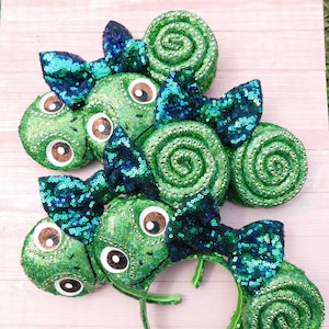 Peut inclure: Plusieurs bandeaux verts à paillettes avec un motif de caméléon. Chaque bandeau présente un grand visage de caméléon rond avec des yeux bruns et un nœud à paillettes dans les tons bleu sarcelle et bleu. Les oreilles sont en spirale.