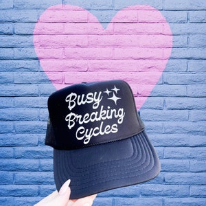 May include: Black trucker hat with the text "Busy Breaking Cycles" in white script and star graphics. The hat has a mesh back and a curved brim. The background features a brick wall with a pink heart.