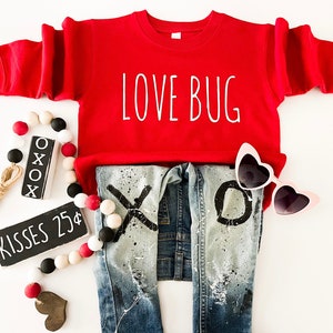 Pode incluir: Um moletom vermelho com o texto "LOVE BUG" em branco, combinado com jeans de ganga desgastados com desenhos "XO". Inclui óculos de sol em forma de coração, uma grinalda de contas e um letreiro que diz "KISSES 25¢".