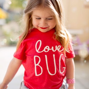 May include: A young child wearing a red t-shirt with the words "love BUG" in white script. The child is also wearing a light blue skirt and a pink bracelet. The child has blonde hair and red lipstick.