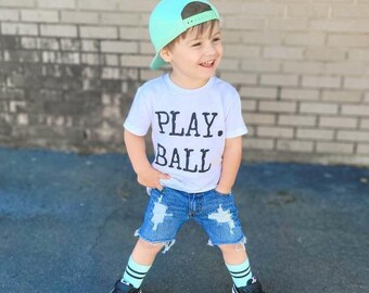 May include: A young child wearing a white t-shirt with the words "PLAY. BALL" in black, paired with distressed denim shorts. The outfit is completed with a teal baseball cap, black sneakers, and teal and black striped socks. The child is smiling.