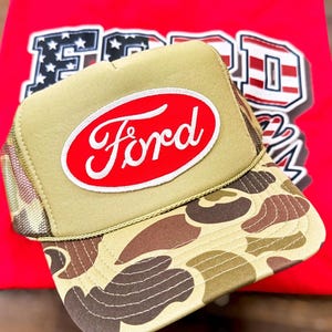 May include: A tan trucker hat with a camouflage brim and a red oval patch featuring the white Ford logo. The hat is positioned over a bright red t-shirt that displays a large graphic with the word Ford in a patriotic American flag pattern.