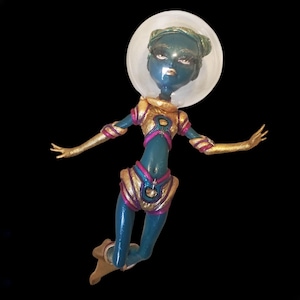 May include: A teal and gold astronaut doll with a clear plastic helmet. The doll has pink accents and is posed with arms outstretched.