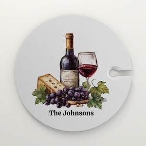 May include: Round white coaster with a watercolor illustration of a bottle of wine, a glass of red wine, a wedge of cheese, and grapes. The text "The Johnsons" is printed below the illustration.