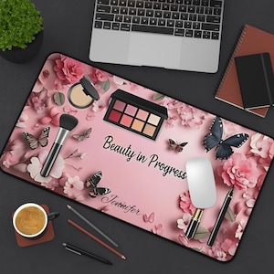 May include: A pink desk mat with a floral and butterfly design, featuring makeup items like a palette, brush, and compact. The text "Beauty in Progress" and a name are printed on the mat. A laptop, notebook, and coffee cup are also visible.