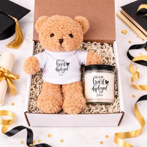 Puede incluir: Un oso de peluche color canela con una camiseta blanca con el texto Half of my heart is deployed John y un tarro de vela de cristal a juego con tapa negra que presenta el mismo texto, ambos en una caja de regalo de cartón con relleno.