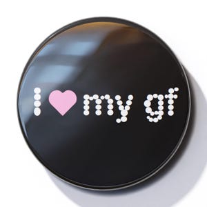 Könnte beinhalten: Ein schwarzer Knopf mit weißen Punkten, die "I love my gf" buchstabieren, mit einem rosa Herzen in der Mitte.