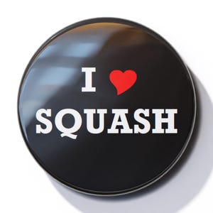 Puede incluir: Botón negro y blanco con el texto "Me encanta el squash" y un corazón rojo.