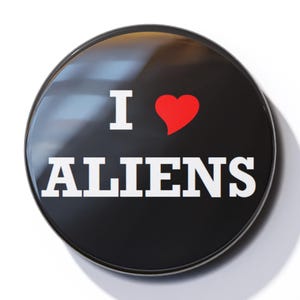 Könnte beinhalten: Ein schwarzer Knopf mit weißem Rand und dem Text "I ♥ ALIENS" in weißen Buchstaben. Das Herz ist rot.