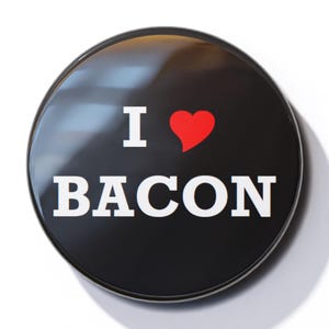 Può includere: Un bottone nero con la scritta "Amo il bacon" in bianco e un cuore rosso.