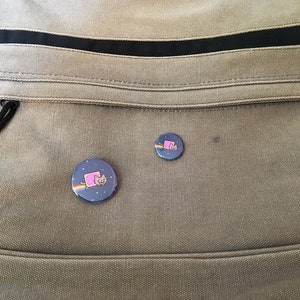 May include: Two round, blue and white buttons with a pink cat wearing a rainbow on a gray canvas bag.