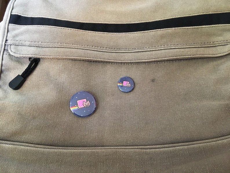 May include: Two round, blue and white buttons with a pink cat wearing a rainbow on a gray canvas bag.