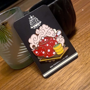 Puede incluir: Una tetera roja y blanca con un patrón de flores blancas, con vapor saliendo del pico, sentada en una bandeja dorada con una taza verde y amarilla. La tetera está sobre un fondo negro con el texto "Moonbrew Atelier" en blanco.