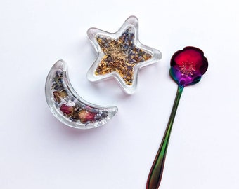 May include: A set of two clear resin dishes shaped like a crescent moon and a star, filled with dried flowers and herbs. A rainbow iridescent metal spoon with a flower design is also included.