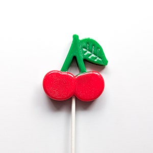 May include: A red and green cherry lollipop on a white stick. The cherry is made of candy and has a shiny finish.