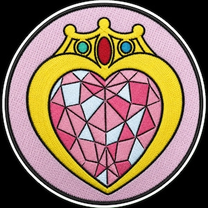 Op de afbeelding: Een ronde geborduurde patch met een hartvormig ontwerp met een gefacetteerd roze en wit kristalpatroon. Het hart is omlijst door een gele rand met een kroonachtige bovenkant, geaccentueerd door een centrale rode edelsteen en twee blauwe edelstenen.