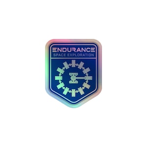 May include: A shield-shaped sticker with a holographic border featuring a dark blue background. The text ENDURANCE SPACE EXPLORATION is printed in white at the top, above a white graphic of a circular space station with a central rotating arm.