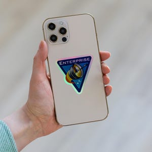 Pode incluir: Um smartphone de cor clara com um autocolante triangular na parte de tr&aacute;s. O autocolante tem um fundo azul, a palavra ENTERPRISE em letras brancas e um gr&aacute;fico de uma antena parab&oacute;lica com um rasto amarelo e laranja.