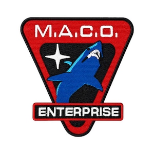 Puede incluir: Un parche bordado triangular con borde rojo y ribete negro. La parte superior presenta el texto blanco M.A.C.O. sobre un campo triangular negro que contiene un gráfico de tiburón azul y una estrella blanca de cuatro puntas. Una pestaña rectangular roja en la parte inferior muestra el texto blanco ENTERPRISE.