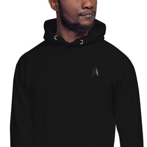May include: Black hooded sweatshirt with a small, gray and black emblem on the chest. The hoodie has a drawstring and a front pocket. The material appears to be a soft, comfortable fabric.