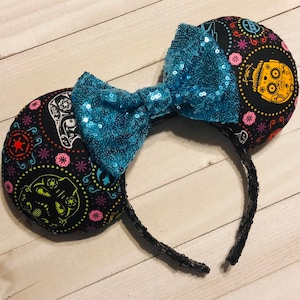 May include: Black mouse ears with a blue sequined bow. The ears are decorated with a colorful Day of the Dead pattern featuring Star Wars characters.