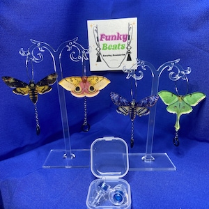 May include: Four pairs of moth-shaped earrings in various colours, displayed on clear stands. The earrings have intricate wing patterns and dangle from hoops. A small clear case with blue earplugs is in front. The sign says "Funky Beats Earplug Accessories."
