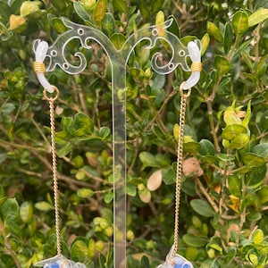 Peut inclure: Boucles d'oreilles en acrylique transparent avec chaîne et détails dorés. Les boucles d'oreilles présentent un design unique avec des éléments bleus et transparents en forme de bouchons d'oreille. Elles sont présentées sur un fond de feuillage vert.