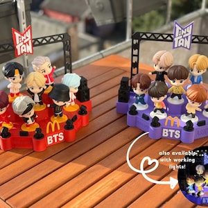 Peut inclure: Deux figurines Tiny Tan BTS sur des plateformes en forme de scène. L'une est rouge, l'autre violette, toutes deux avec le logo McDonald's et le texte BTS. Chaque plateforme contient sept figurines aux couleurs de cheveux différentes. Le texte "Tiny Tan" est en haut.