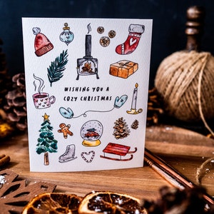 May include: A white Christmas card with a watercolor illustration of various winter holiday icons, including a fireplace, a Christmas tree, a stocking, a snow globe, and a gingerbread man. The text "Wishing you a Cozy Christmas" is written in black lettering.