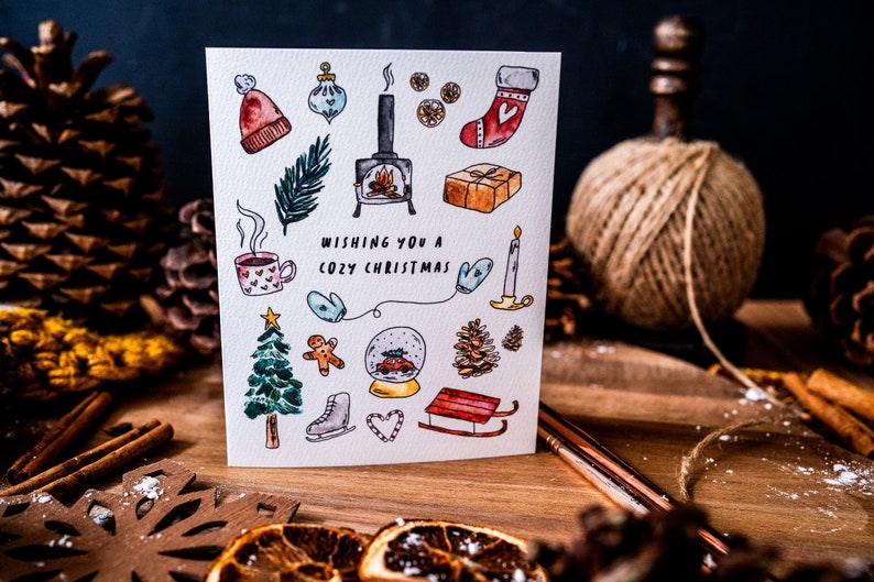 May include: A white Christmas card with a watercolor illustration of various winter holiday icons, including a fireplace, a Christmas tree, a stocking, a snow globe, and a gingerbread man. The text "Wishing you a Cozy Christmas" is written in black lettering.