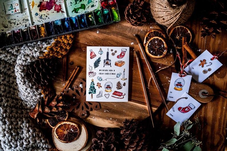 May include: A white card with a hand-drawn illustration of winter holiday themed items, including a fireplace, a Christmas tree, a gingerbread man, a snow globe, and a stocking. The card reads "Wishing you a cozy Christmas". The card is surrounded by various craft supplies, including pine cones, cinnamon sticks, dried orange slices, and a ball of twine.