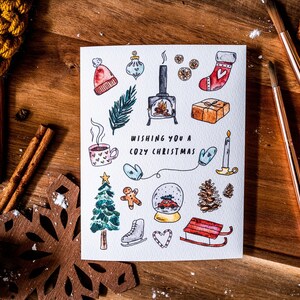 May include: A white card with a hand-drawn illustration of winter holiday themed items, including a fireplace, a Christmas stocking, a gingerbread man, a snow globe, and a Christmas tree. The text "Wishing you a cozy Christmas" is written in black lettering.