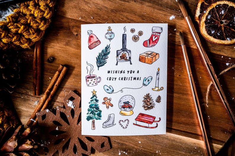 May include: A white card with a hand-drawn illustration of winter holiday themed items, including a fireplace, a Christmas stocking, a gingerbread man, a snow globe, and a Christmas tree. The text "Wishing you a cozy Christmas" is written in black lettering.