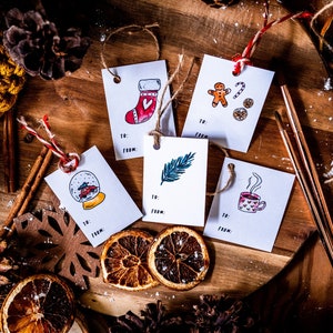 May include: Five white gift tags with different illustrations on a wooden surface. The illustrations include a stocking, a gingerbread man, a feather, a snow globe, and a cup of hot chocolate. The tags are tied with twine and have the words "To:" and "From:" printed on them.