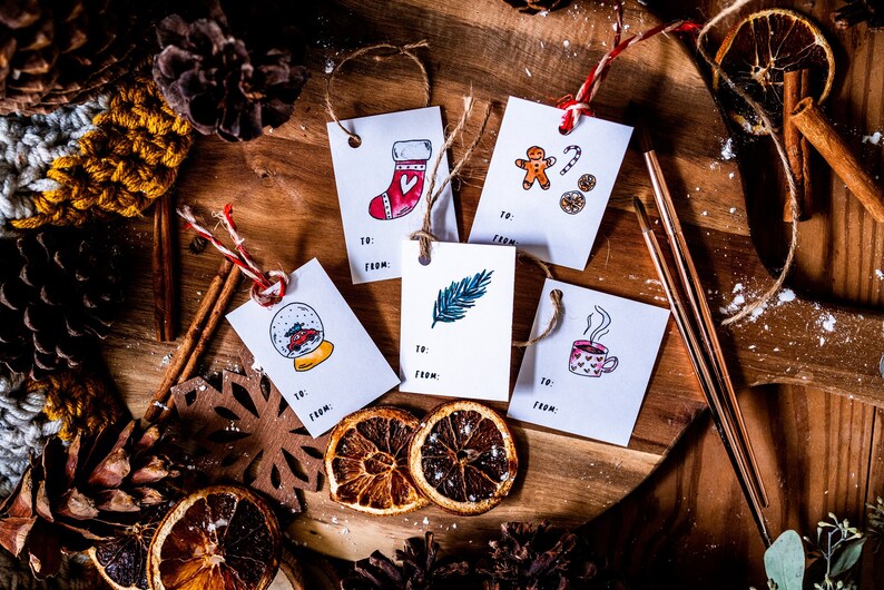 May include: Five white gift tags with different illustrations on a wooden surface. The illustrations include a stocking, a gingerbread man, a feather, a snow globe, and a cup of hot chocolate. The tags are tied with twine and have the words "To:" and "From:" printed on them.