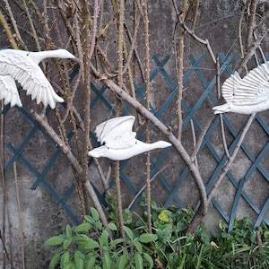 May include: Three white, decorative goose sculptures in flight are mounted on a weathered gray wall. The geese are positioned at different heights, with wings spread, and are set against a backdrop of bare branches and a green lattice.