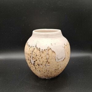 Può includere: Un piccolo vaso rotondo in ceramica con un'ampia apertura. Il vaso ha una parte superiore color crema e un corpo strutturato color marrone chiaro con un aspetto invecchiato e usurato. Il vaso è adatto per esporre fiori o come elemento decorativo.