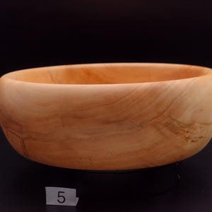 May include: A handcrafted wooden bowl with a natural, light brown finish. The bowl has a smooth, rounded shape and visible wood grain patterns. The number "5" is visible on a small white label. This bowl is suitable for serving or display.