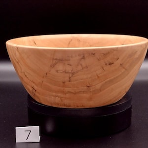 May include: A handcrafted wooden bowl with a light brown color and visible wood grain. The bowl has a wide, open design and sits on a black base. The number "7" is visible on a small white label.
