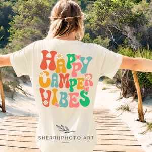 May include: A white t-shirt with a retro-style graphic that says "Happy Camper Vibes" in colorful, wavy letters with flowers and butterflies. The shirt is being worn by a person standing on a wooden walkway.