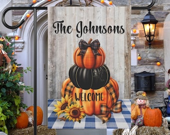 May include: A fall-themed welcome garden flag with a stack of three pumpkins, the top one with a brown bow. The flag has a wooden background with the text "The Johnsons" and "Welcome" in black lettering. The flag is displayed on a blue and white checkered background.
