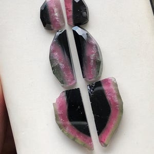 May include: Six pieces of watermelon tourmaline gemstone cabochons in various shades of pink, green, and black. The cabochons are cut in a variety of shapes, including ovals and half-ovals.