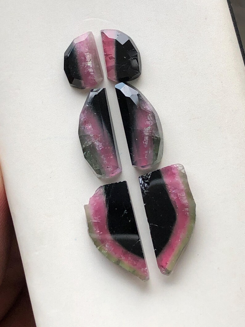 May include: Six pieces of watermelon tourmaline gemstone cabochons in various shades of pink, green, and black. The cabochons are cut in a variety of shapes, including ovals and half-ovals.